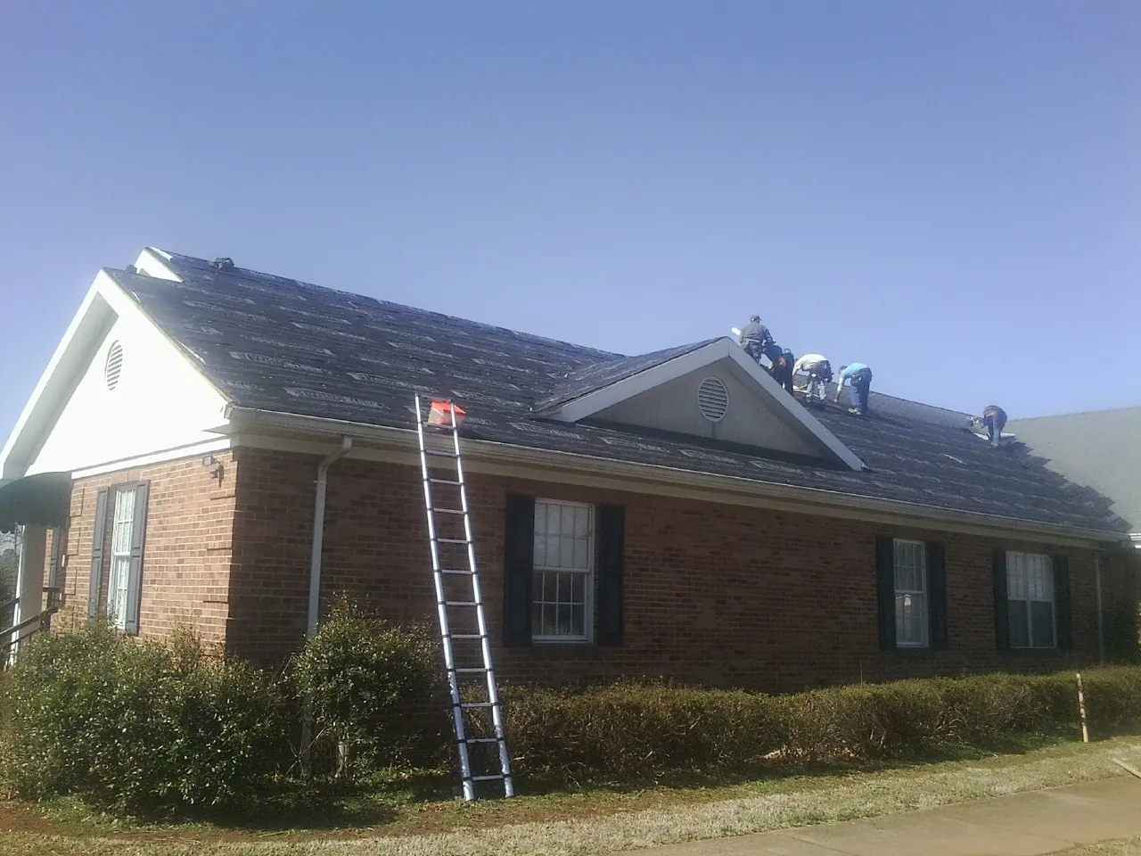 Crew tearing shingles off a brick ranch roof with a ladder set — Emergency Roof Repair work by our crew in Day