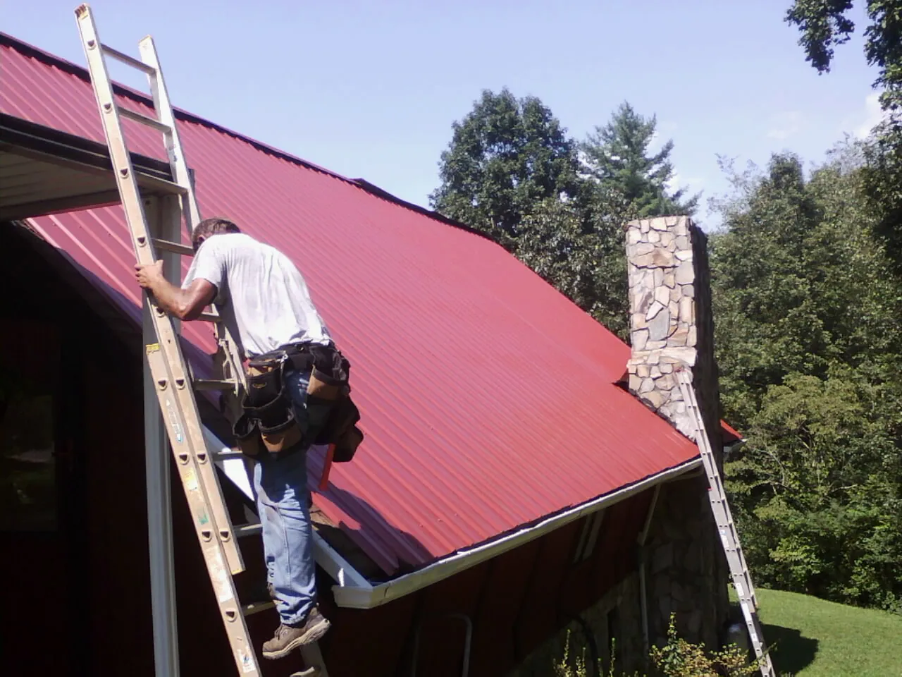 Roofer on a ladder beside a red metal roof and stone chimney — Soffit & Fascia Repair by Roofing Brothers in Day